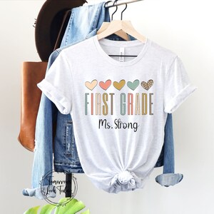 May include: White t-shirt with the text "FIRST GRADE Ms. Strong" in colorful letters. A row of five hearts in different colors and patterns are above the text.