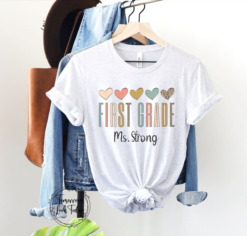 May include: White t-shirt with the text "FIRST GRADE Ms. Strong" in colorful letters. A row of five hearts in different colors and patterns are above the text.