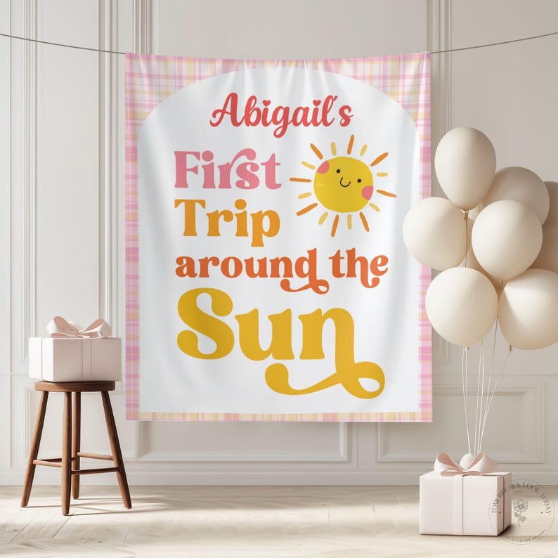 May include: A banner with the text "Abigail's First Trip around the Sun" in pink, orange, and yellow. A smiling sun graphic is included. The banner is framed with a pink plaid pattern. Balloons and a gift box are also in the image.