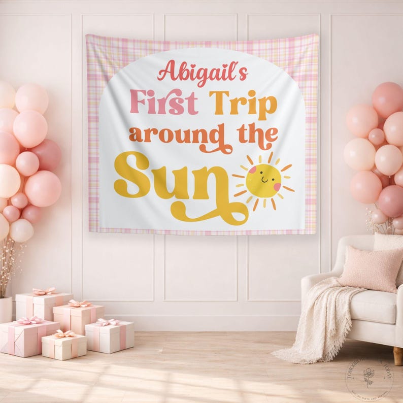 May include: A white banner with the text "Abigail's First Trip around the Sun" in pink, orange, and yellow. The banner has a smiling sun graphic and is set against a pink plaid background. Balloons and gifts are in the foreground.