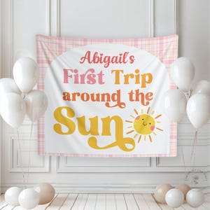 May include: A party backdrop with the text "Abigail's First Trip around the Sun" in pink and orange, with a smiling sun illustration. The backdrop is framed by a pink plaid pattern and surrounded by white and beige balloons.