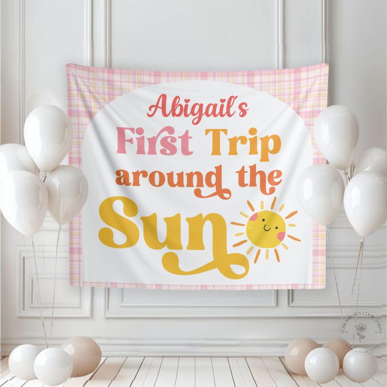 May include: A party backdrop with the text "Abigail's First Trip around the Sun" in pink and orange, with a smiling sun illustration. The backdrop is framed by a pink plaid pattern and surrounded by white and beige balloons.