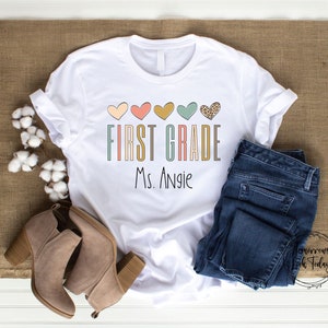 May include: White t-shirt with a colorful graphic that says "First Grade" with hearts and the name "Ms. Angie" printed below.