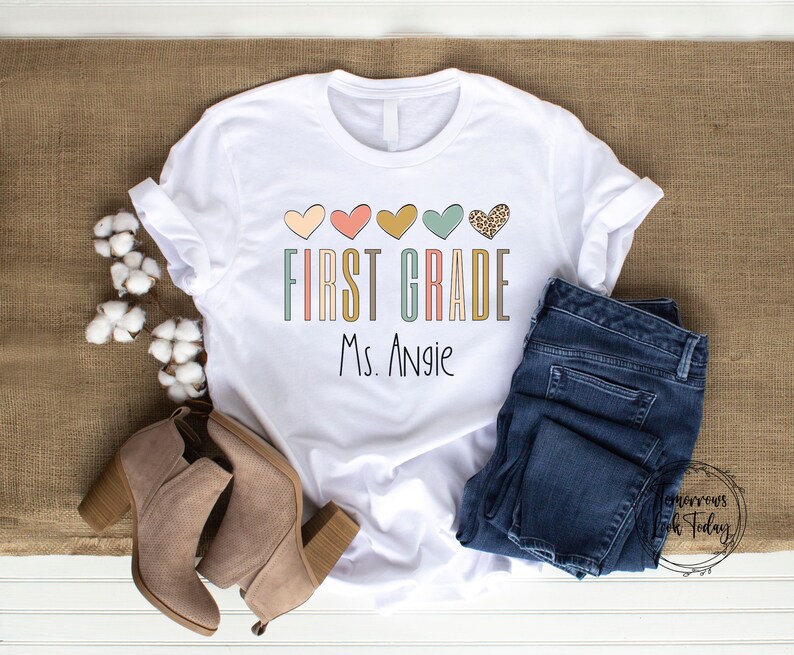 May include: White t-shirt with a colorful graphic that says "First Grade" with hearts and the name "Ms. Angie" printed below.