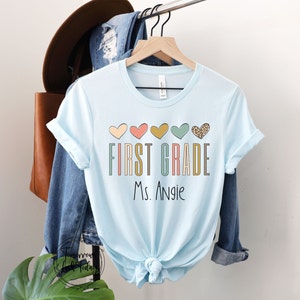 May include: Light blue t-shirt with a graphic design of colorful hearts and the text "FIRST GRADE Ms. Angie".