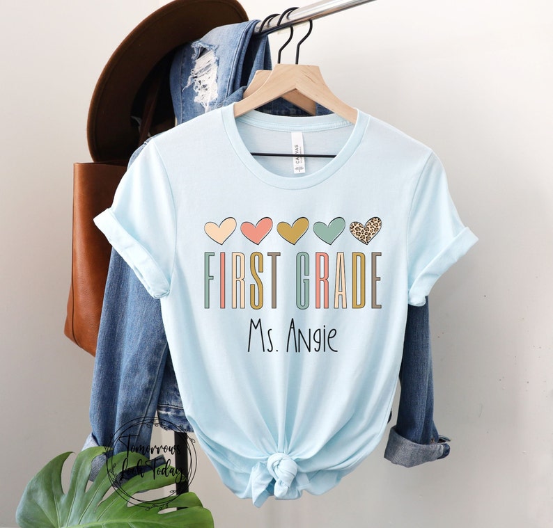 May include: Light blue t-shirt with a graphic design of colorful hearts and the text "FIRST GRADE Ms. Angie".