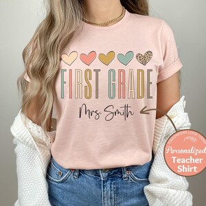May include: Pink t-shirt with the text "First Grade" in colorful letters and a row of colorful hearts above the text. The text "Mrs. Smith" is below the text "First Grade" with an arrow pointing to the right.