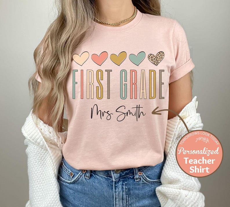 May include: Pink t-shirt with the text "First Grade" in colorful letters and a row of colorful hearts above the text. The text "Mrs. Smith" is below the text "First Grade" with an arrow pointing to the right.