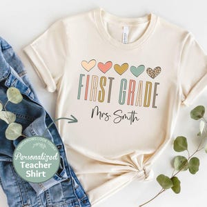 May include: A beige t-shirt with a row of colorful hearts and the text "FIRST GRADE" in a colorful font. The text "Mrs. Smith" is written below the word "GRADE".