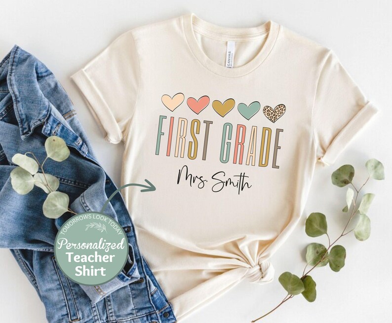 May include: A beige t-shirt with a row of colorful hearts and the text "FIRST GRADE" in a colorful font. The text "Mrs. Smith" is written below the word "GRADE".