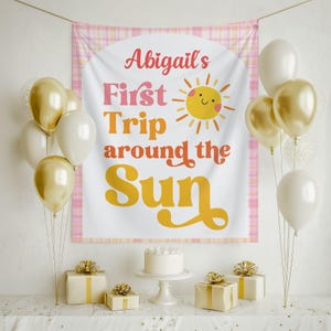 May include: A party scene with a banner that reads "Abigail's First Trip around the Sun" with a sun illustration. Gold and white balloons, wrapped gifts, and a cake on a stand are arranged on a table, creating a festive atmosphere.