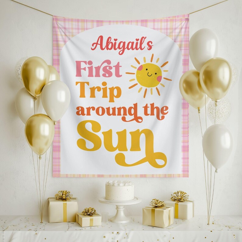 May include: A party scene with a banner that reads "Abigail's First Trip around the Sun" with a sun illustration. Gold and white balloons, wrapped gifts, and a cake on a stand are arranged on a table, creating a festive atmosphere.