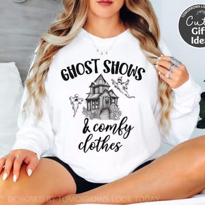 May include: White sweatshirt with black text that reads "Ghost Shows & Comfy Clothes". The text is surrounded by a drawing of a haunted house and ghosts.