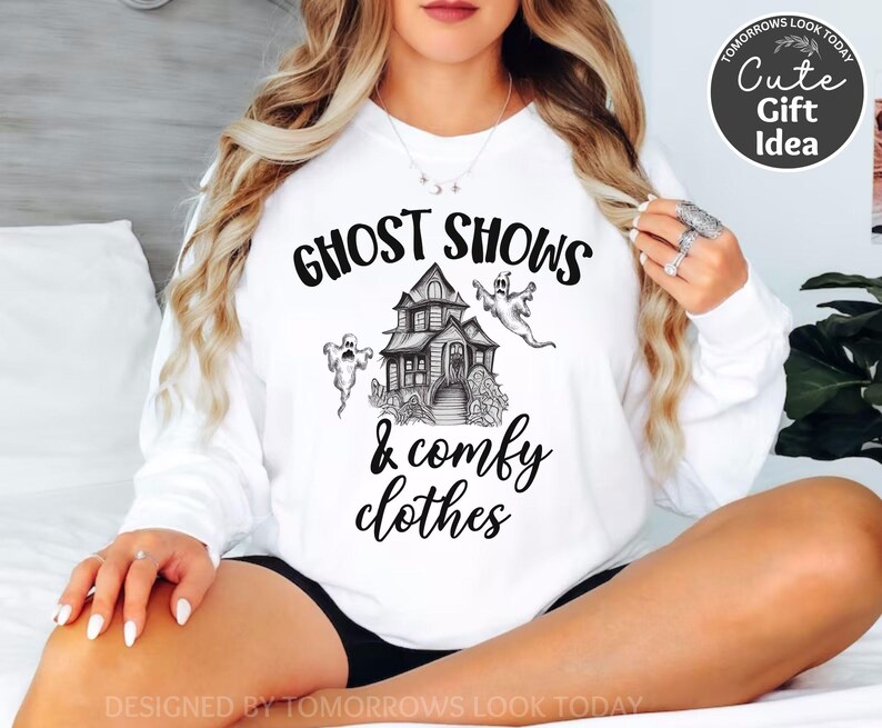 May include: White sweatshirt with black text that reads "Ghost Shows & Comfy Clothes". The text is surrounded by a drawing of a haunted house and ghosts.