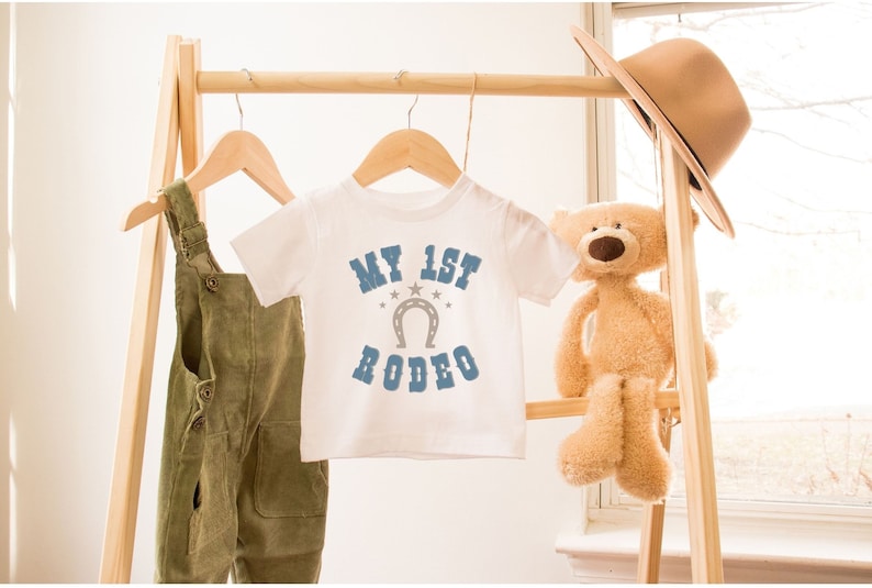 May include: White baby onesie with blue text that says "My 1st Rodeo" with a horseshoe and stars. The onesie is hanging on a wooden rack with a teddy bear and a brown hat.
