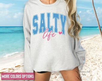May include: A light gray sweatshirt with the words "SALTY life" in blue and pink lettering. The word "SALTY" is in a block letter style, and the word "life" is in a cursive font. A pink heart is next to the word "life". The sweatshirt is displayed on a beach.