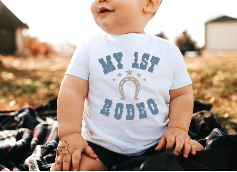 May include: A white baby onesie with blue text that reads "My 1st Rodeo" with a brown horseshoe and stars.