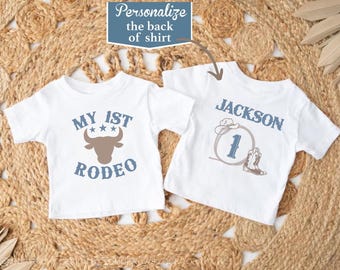 May include: Two white short-sleeve t-shirts. The left shirt features a brown bull head graphic with the text "MY 1ST RODEO" in blue. The right shirt displays the name "JACKSON" in blue above a lasso, a cowboy hat, a number one, and a boot.