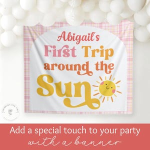 May include: A white banner with the text "Abigail's First Trip around the Sun" in pink and yellow. A smiling sun graphic is on the right. The banner has a pink plaid border and is decorated with white balloons.