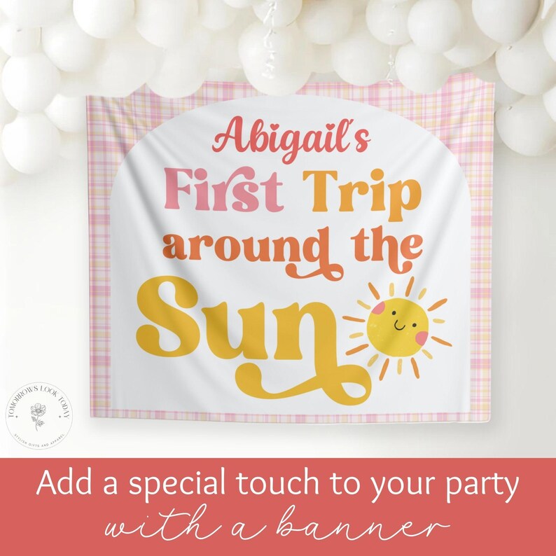 May include: A white banner with the text "Abigail's First Trip around the Sun" in pink and yellow. A smiling sun graphic is on the right. The banner has a pink plaid border and is decorated with white balloons.