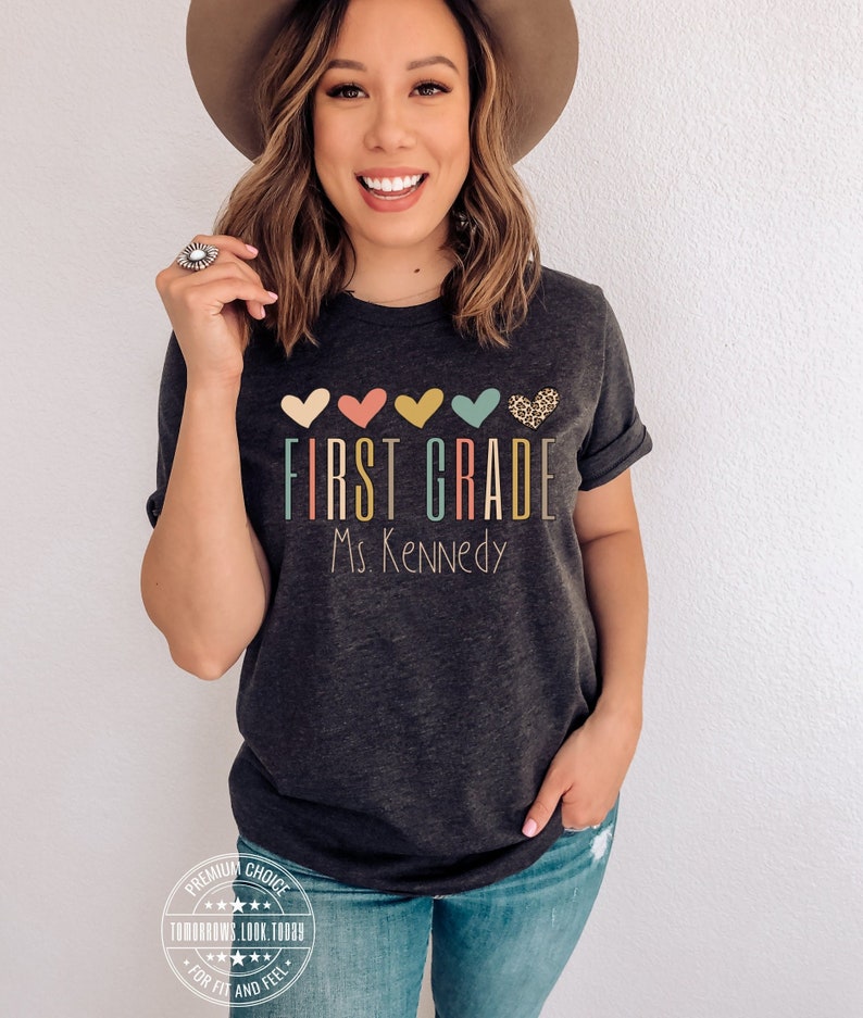 May include: A dark gray t-shirt with the text "FIRST GRADE" in colorful letters and "Ms. Kennedy" below. The text is surrounded by five colorful hearts, one of which has a leopard print pattern.
