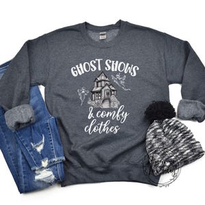 May include: A dark gray sweatshirt with the text "Ghost Shows & Comfy Clothes" in white. The design features a cartoon haunted house with ghosts flying around it.