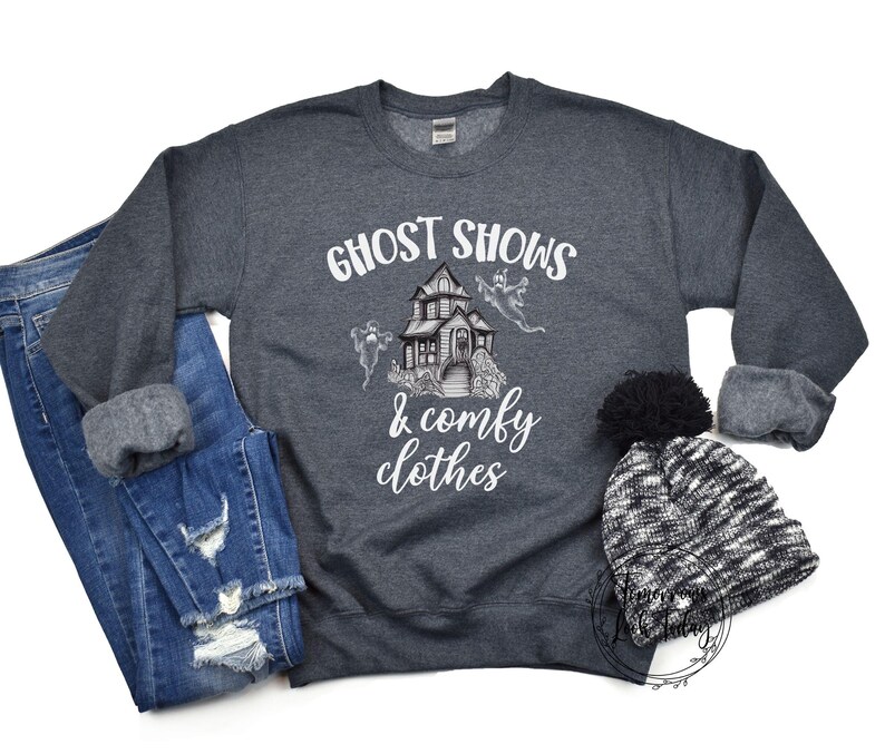 May include: A dark gray sweatshirt with the text "Ghost Shows & Comfy Clothes" in white. The design features a cartoon haunted house with ghosts flying around it.