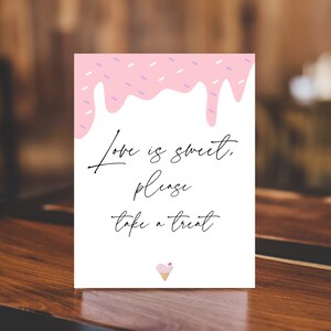 May include: A white sign with pink dripping icing on top. The sign says "Love is sweet, please take a treat" with a pink ice cream cone icon at the bottom.