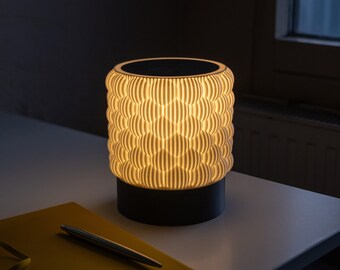 May include: A white table lamp with a geometric pattern on the shade. The lamp is turned on and casting a warm glow. The lamp is sitting on a white table next to a yellow notebook and a pen.