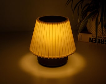 May include: A small, portable table lamp with a pleated, off-white conical shade that emits a warm, soft yellow glow. The lamp features a dark, cylindrical base and is illuminated from within, casting a circular pool of light beneath it.