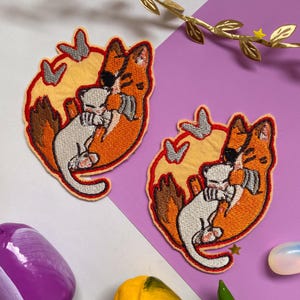 May include: Two embroidered patches featuring a cartoon fox with an eye patch hugging a white creature. The patches are orange, yellow, and white, with a red border. The patches are on a white and purple background.