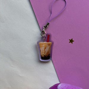 Peut inclure: Une breloque en acrylique transparent en forme de tasse de thé aux perles avec une paille rouge, remplie d'un liquide beige et de perles de tapioca brun foncé au fond. La breloque est fixée à un cordon élastique violet clair avec un fermoir mousqueton.