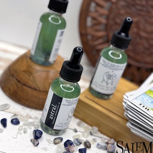 May include: Three glass bottles containing green liquid with black droppers. The bottles are labelled 'astral' and feature text about 'travel the astral' and 'spiritual connection'. The bottles are positioned on a wooden surface surrounded by small blue and white stones.