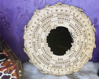May include: A wooden circular divination board with a mirror in the center. The board is decorated with floral designs and features a calendar, days of the week, months of the year, and the alphabet. The board is on a white furry surface.