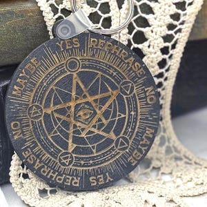May include: A black and gold round keychain with the words "YES REPHRASE NO MAYBE" around the edge. The center features a star and eye design. The keychain is on a white lace doily with green and purple stones. The text "SALEM SUPPLY CO" is visible.