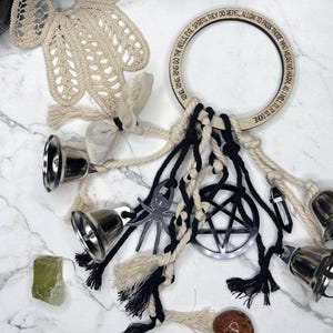 May include: A collection of spiritual items including a wooden ring with engraved text, a pentagram charm, silver bells, crystals, and a crocheted hand. The items are arranged on a marble surface.