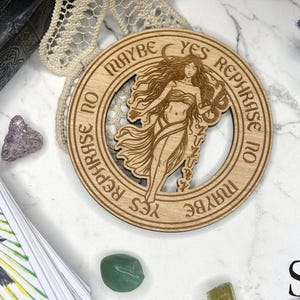 May include: A wooden divination board with a woman figure in the center, surrounded by the words "Maybe, Yes, Rephrase, No". The image includes tarot cards, crystals, and a moon-shaped charm. The Salem Ply Co. logo is visible.