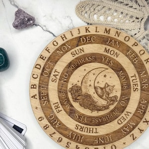 May include: A wooden planchette with engraved letters, numbers, and a crescent moon design with a cat. The planchette is surrounded by crystals, tarot cards, and the text "SALEM Co. Blessed be the Witches."