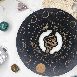 May include: A black and gold planchette with a snake design in the center, surrounded by letters, numbers, and crescent moons. The planchette has the words "yes", "no", and "goodbye" on it. The Salem Supply Co. logo is visible.