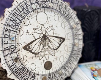 May include: A round, wooden planchette with a moth illustration and the words "YES", "NO", "MAYBE", and "UNCLEAR". The planchette has a decorative edge and is surrounded by crystals and a deck of tarot cards. The text "SALEM SUPPLY CO" is visible.