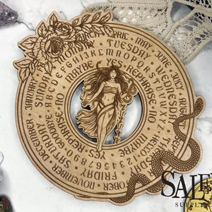 May include: A wooden divination board with a floral and snake design. The board features a central illustration of a woman and includes text such as "Yes", "Maybe", and the days of the week. The board is surrounded by crystals and tarot cards.