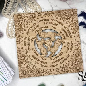 May include: A wooden divination board with a floral border and a central design of three rabbits. The board features engraved text including months, days, and phrases like "YES" and "MAYBE". A deck of tarot cards is visible in the lower left.