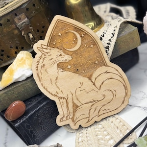 May include: A wooden fox-shaped decoration with a crescent moon and stars design. The fox is sitting, with its tail curled. The background is a light brown color. The text "SALEM SUPPLY CO. BLESSED BE THE WITCHES" is at the bottom.