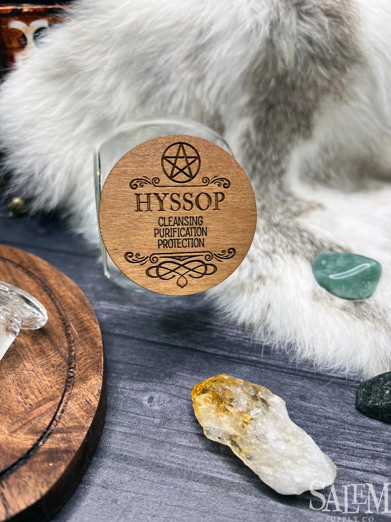 May include: A wooden lid for a glass jar with the word "Hyssop" and the text "Cleansing, Purification, Protection" engraved on it. The lid is decorated with a pentagram and a decorative border.