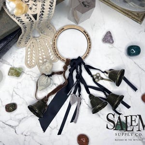 May include: A wooden ring with text, black ribbons, and bronze bells. The ring has the text "PASS THOSE WHO MEAN NO HARM, AS I WILL IT, IT IS DONE" and is surrounded by crystals, a key, and a small axe charm. The Salem Supply Co. logo is in the bottom right.