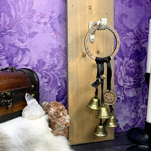 May include: A wooden ring with a black ribbon and bells hanging from it. The ring has the words "The Bells of the Wild Hunt" engraved on it. The ring is attached to a wooden board with a metal hook. There is a small glass bottle with black stones inside hanging from the ribbon. There is a pentagram charm on the ribbon.
