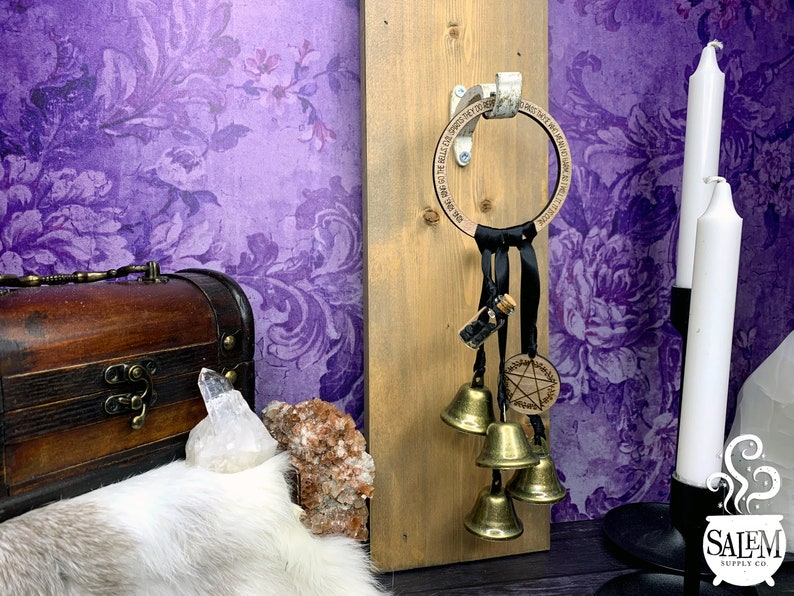 May include: A wooden ring with a black ribbon and bells hanging from it. The ring has the words "The Bells of the Wild Hunt" engraved on it. The ring is attached to a wooden board with a metal hook. There is a small glass bottle with black stones inside hanging from the ribbon. There is a pentagram charm on the ribbon.