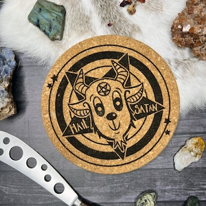 May include: A round cork trivet with a black and white illustration of a cartoon goat head with horns and a pentagram. The goat head is surrounded by a circle with the words "Hail Satan" and stars.
