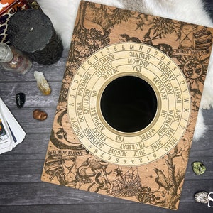 May include: A wooden Ouija board with a black planchette and an intricate design featuring a spider web, a skull, and a raven. The board has a circular alphabet, numbers, and words like "yes", "no", and "goodbye".