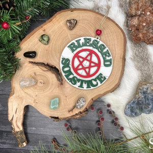 May include: A white wooden ornament with the words "Blessed Solstice" written in green letters around a red pentagram. The ornament is hanging from a wooden slice with various crystals and stones.